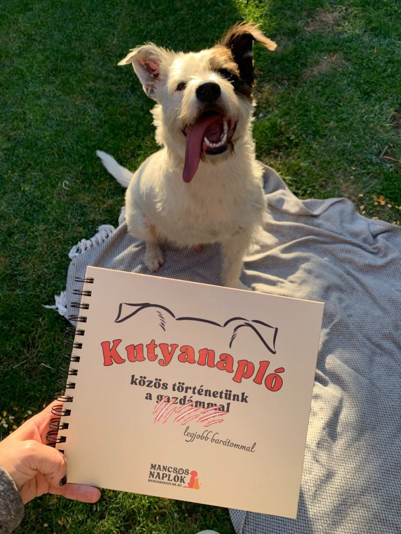 Kutyanapló a Mancsos Naplók sorozat részeként tartva egy női kézben, mögötte egy háromlábú kutya ül kilógó nyelvvel.